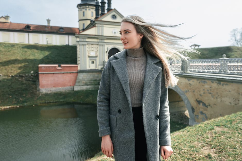 Elegant woman in gray coat near Niasviž Castle on a breezy day.