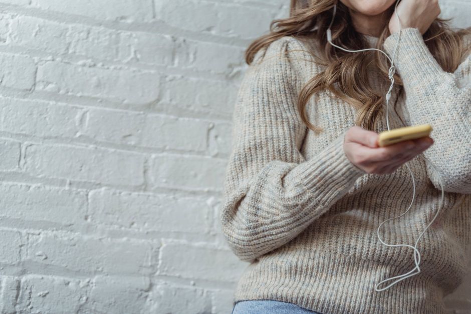 Crop unrecognizable female with long wavy hair in warm sweater listening to music in earphones via mobile phone while sitting near white brick wall