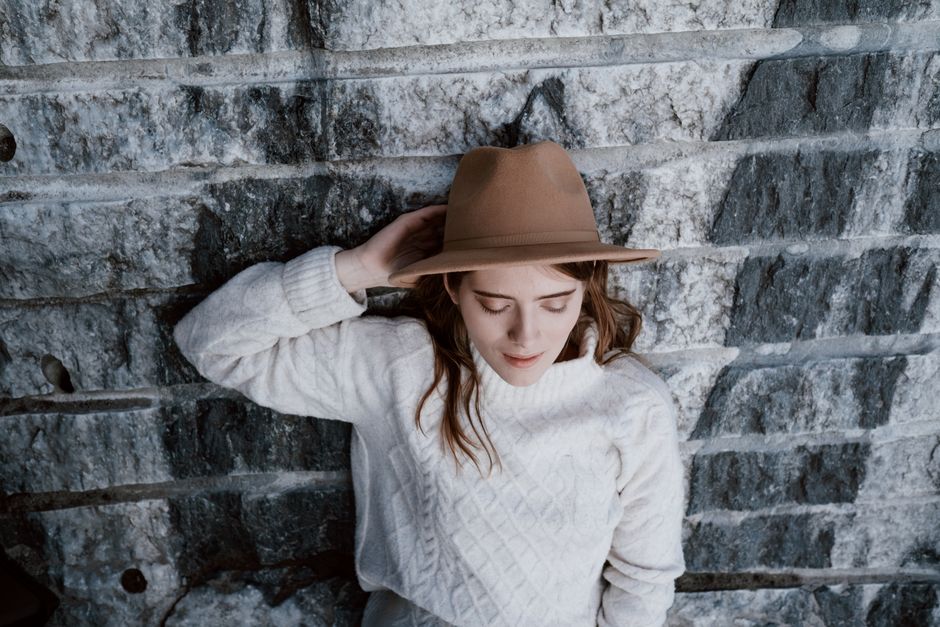 Stylish woman in knit sweater and brown hat, leaning on stone wall.