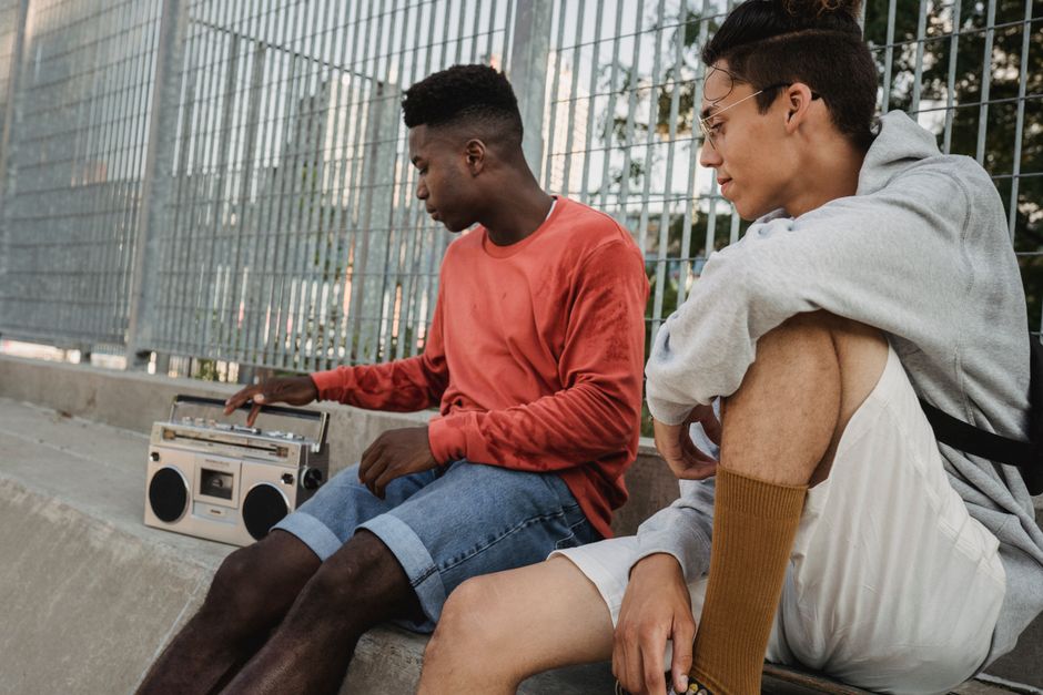 Confident diverse friends chilling together with old fashioned record player while sitting near metal fence of city park on summer day