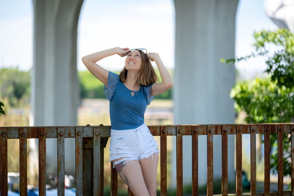 Smiling woman in casual wear enjoying a sunny day outdoors near a rusted fence.