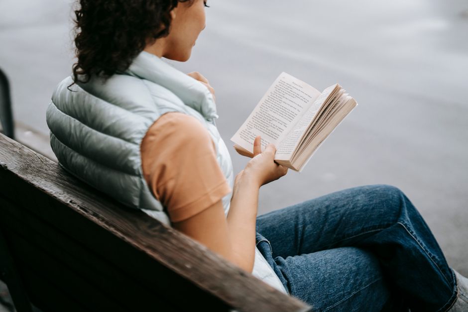 Side view of crop anonymous young female in casual clothes reading interesting book while resting on bench in park