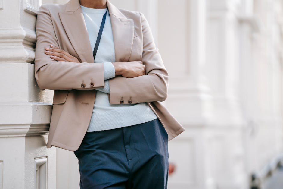 Crop anonymous stylish female manager in smart casual outfit leaning on elegant building wall with folded arms while standing on street on sunny day