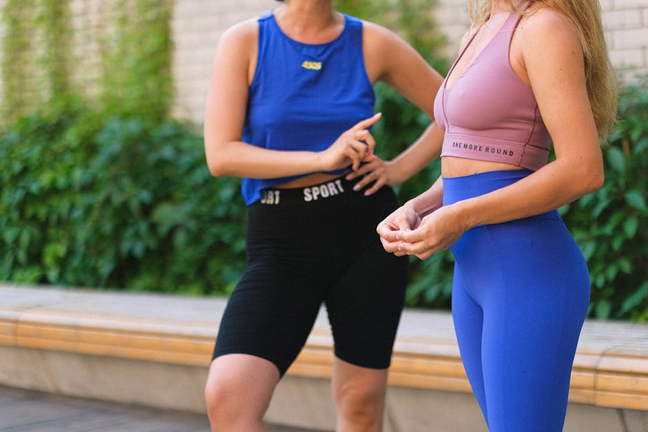 Crop faceless female friends in sportswear standing and talking in street near brick wall with green plants after training in daytime
