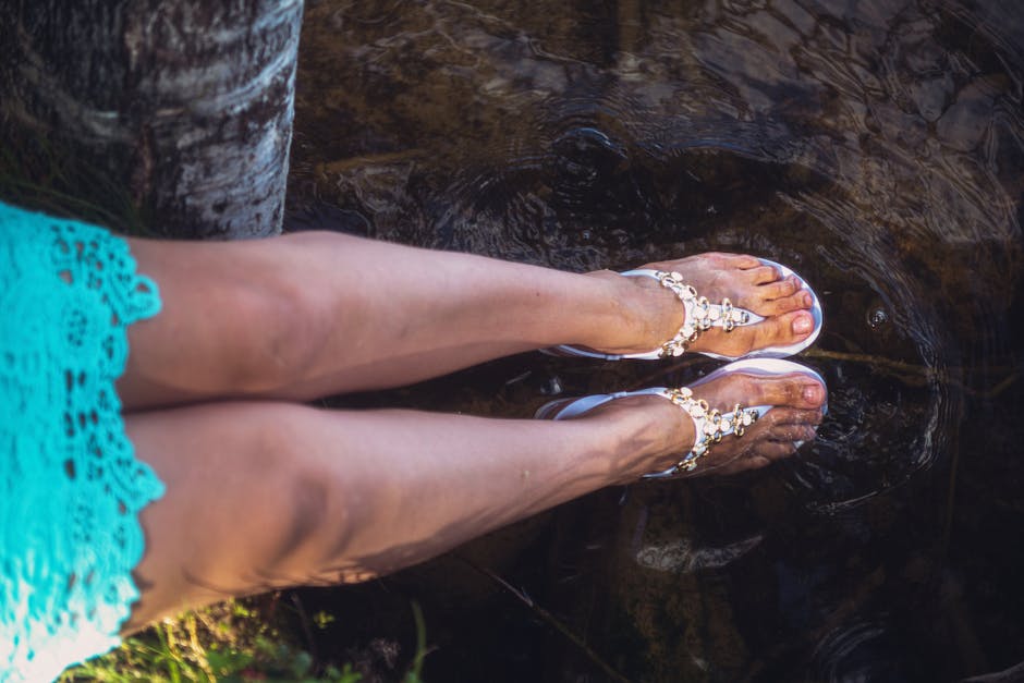 A woman sits by clear water, dipping her feet in, wearing elegant sandals.