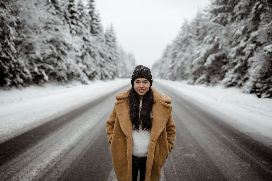 Positive ethnic female in hat and coat standing with hands in pockets on roadway among fir trees covered with snow