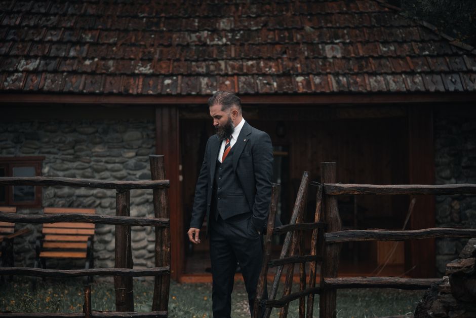 Bearded man in a suit outside a rustic stone cottage with wooden fence.