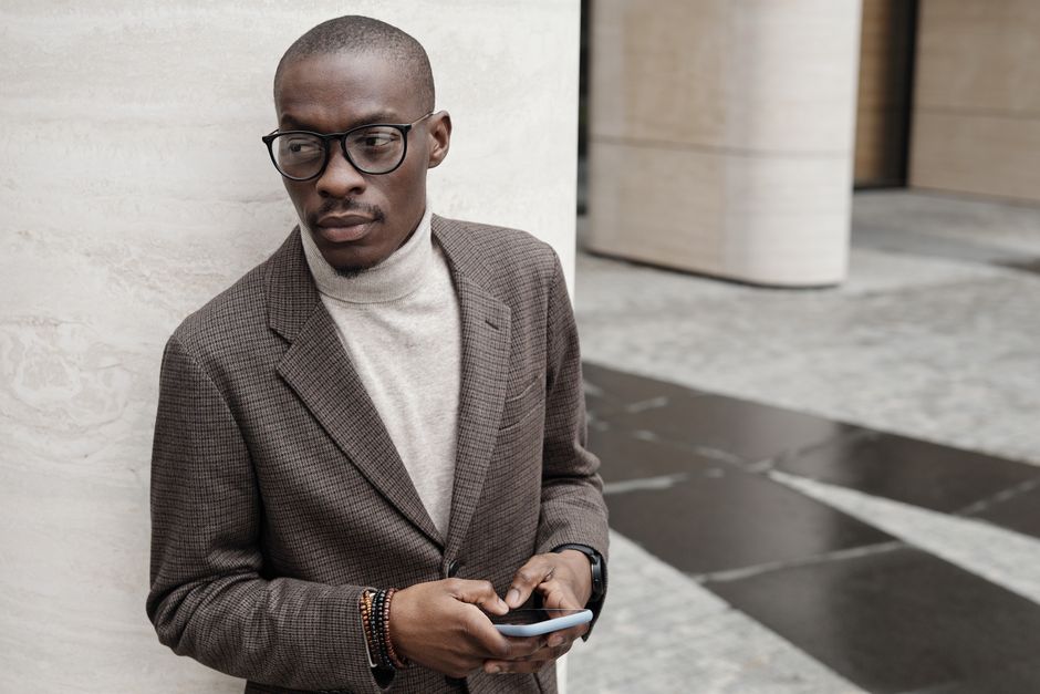 Stylish man in glasses and suit holding a smartphone, leaning against an urban wall.