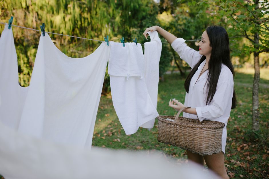 A woman in a white shirt hangs towels on a clothesline in a lush garden setting.