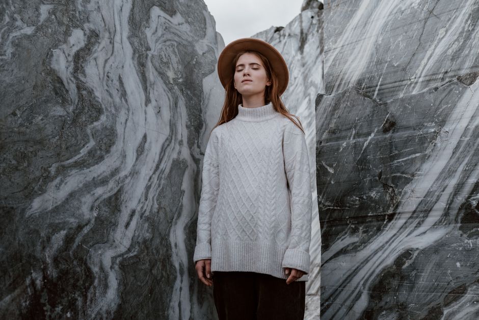 A woman with eyes closed, wearing a wool sweater and fedora, stands between marble walls.