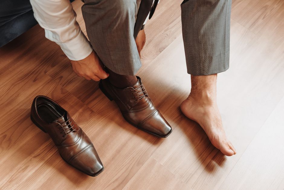 Close-up of a man in a suit putting on brown leather dress shoes indoors on a wooden floor.