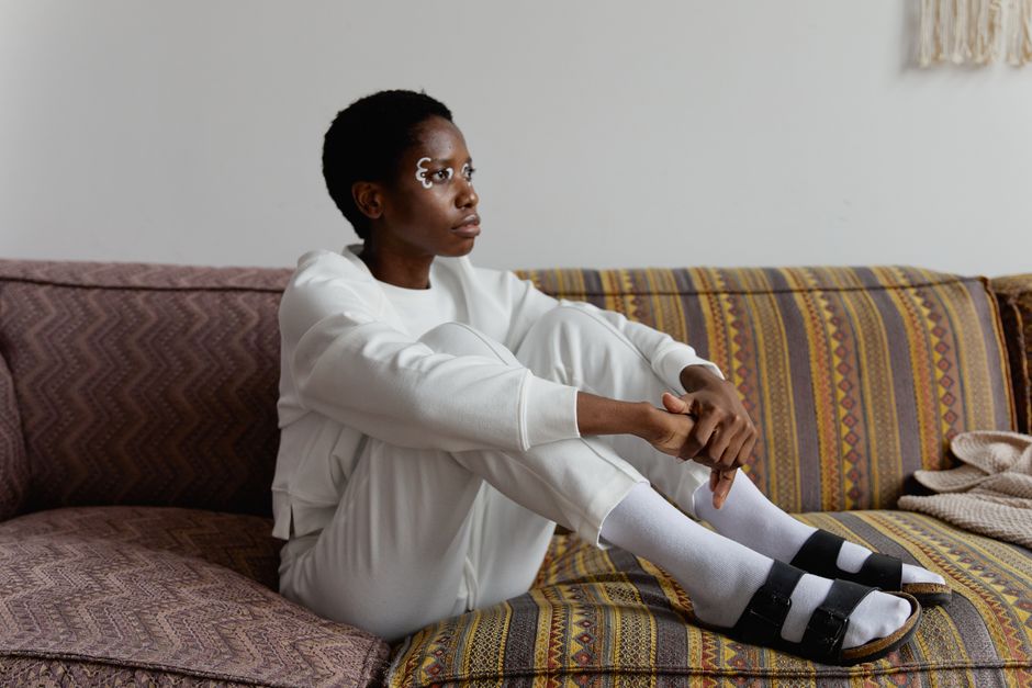 A woman with face paint in white attire, sitting thoughtfully on a patterned sofa.