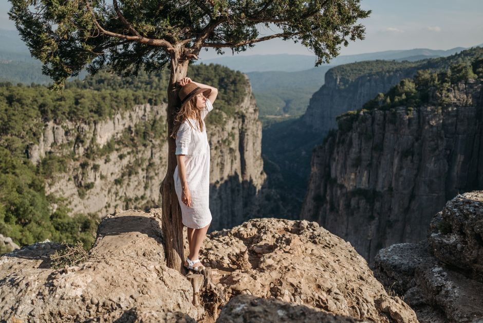 A woman in a white dress and hat stands on a cliff edge amidst scenic mountains in sunlight.