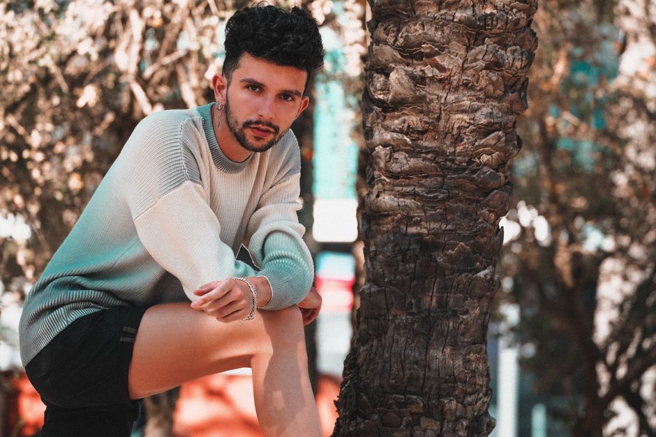 Modern queer male with earring and trendy hairstyle looking at camera leaning on knee in exotic park