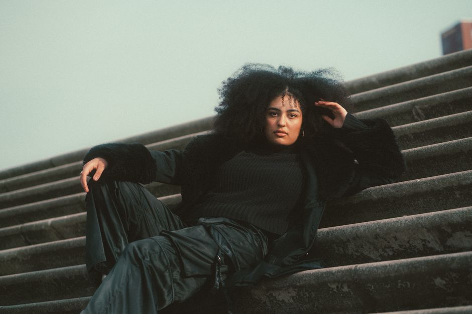 Fashionable woman with afro hairstyle posing confidently on stairs in a trendy outfit.