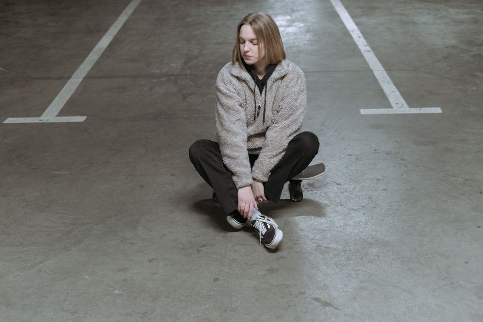 A young woman in casual wear sits on her skateboard in an indoor parking area.