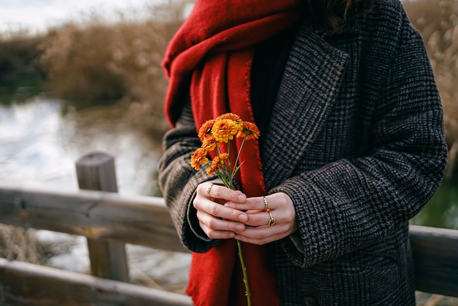 A woman in a coat and red scarf holding orange flowers on a wooden bridge outdoors.