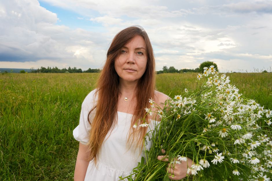 A serene portrait of a woman holding daisies in a green field under a cloudy sky.