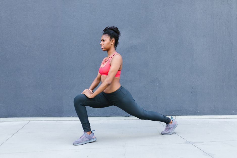 A woman in athletic wear performs a lunge stretch against a gray wall outside, focusing on fitness.