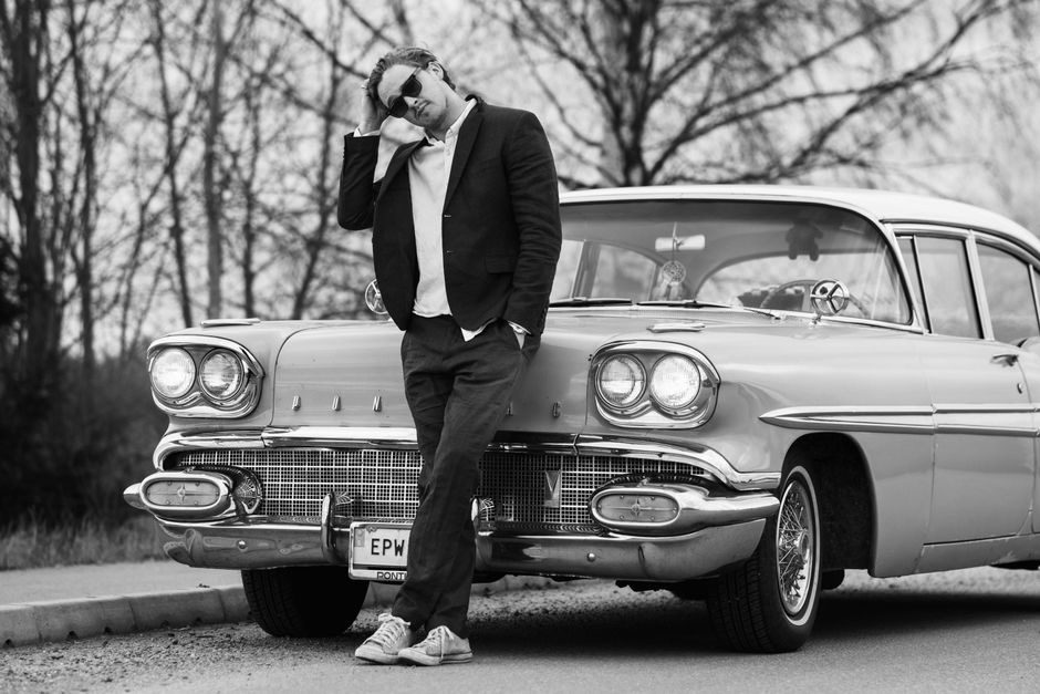 A man in sunglasses poses with a classic Pontiac car on a road.
