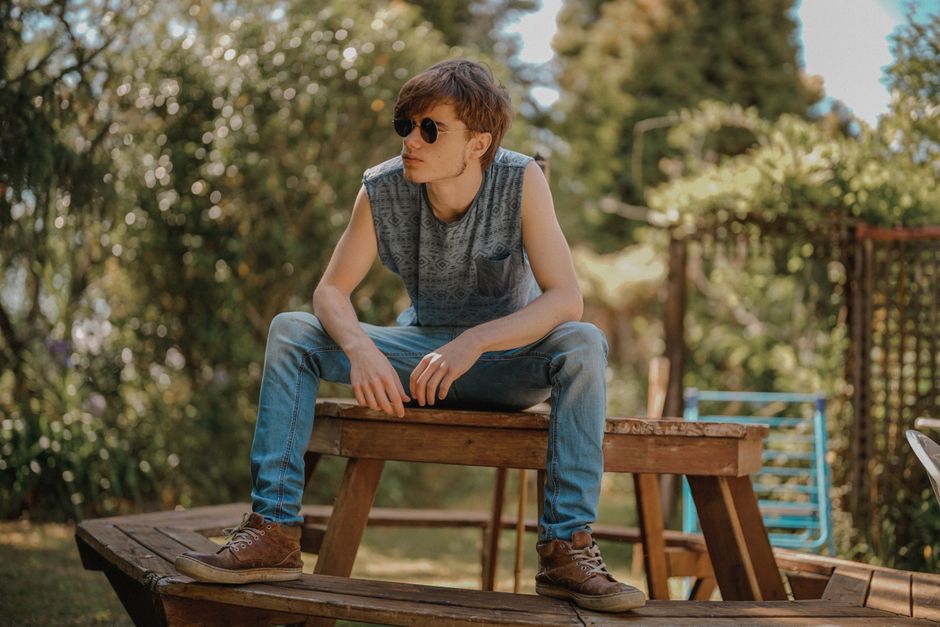 A young man in sunglasses relaxes on a bench outdoors, showcasing a casual summer style.