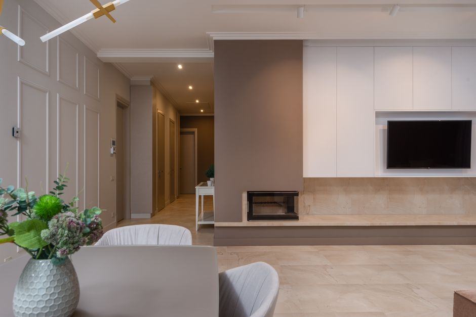 Stylish interior of modern apartment with living room with dining table decorated with potted flowers and big TV set hanging in cabinet opening passing into corridor with several doors