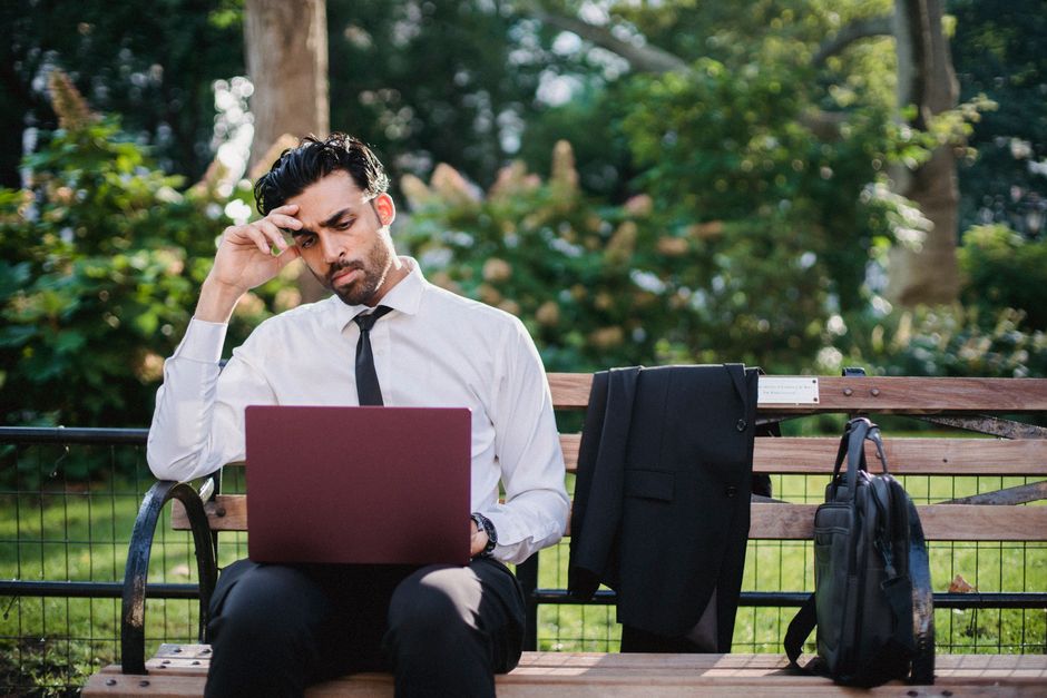 A thoughtful businessman works on his laptop while sitting on a park bench with his coat and bag.