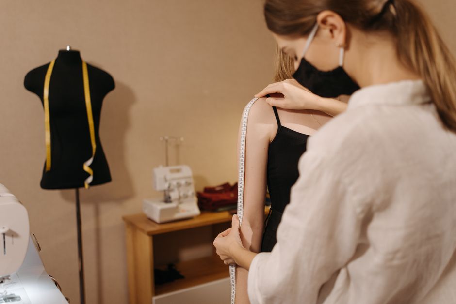 Fashion designer measuring client's shoulder in studio, emphasizing new normal with masks.