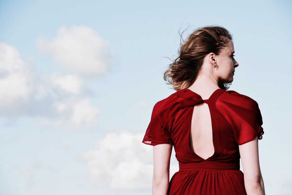 Elegant woman in red dress facing blue sky with clouds, exuding style and grace.