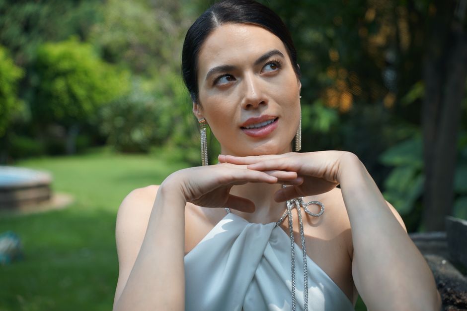 A woman posing outdoors, exuding elegance in a white dress with lush garden surroundings.