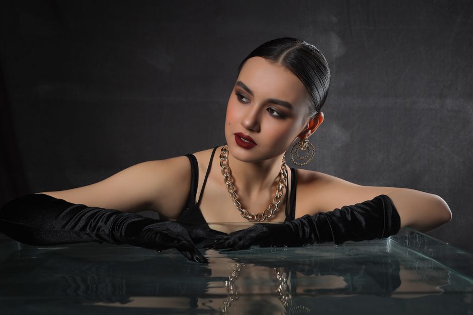 Elegant woman in black attire with bold makeup and jewelry posing against a dark backdrop.