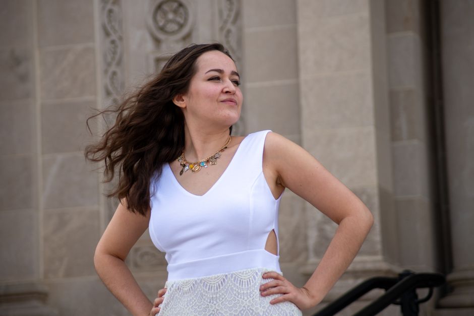 A young woman in a white dress poses confidently in front of an ornate historic building.