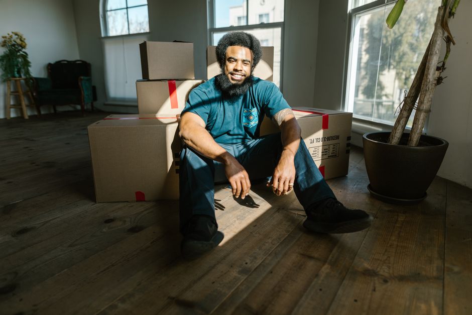 Smiling man sits among moving boxes in a sunny new home, ready for a fresh start.
