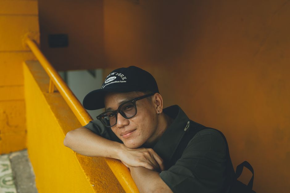 A serene portrait of a man in a baseball cap and glasses leaning on a yellow railing in Ho Chi Minh City.