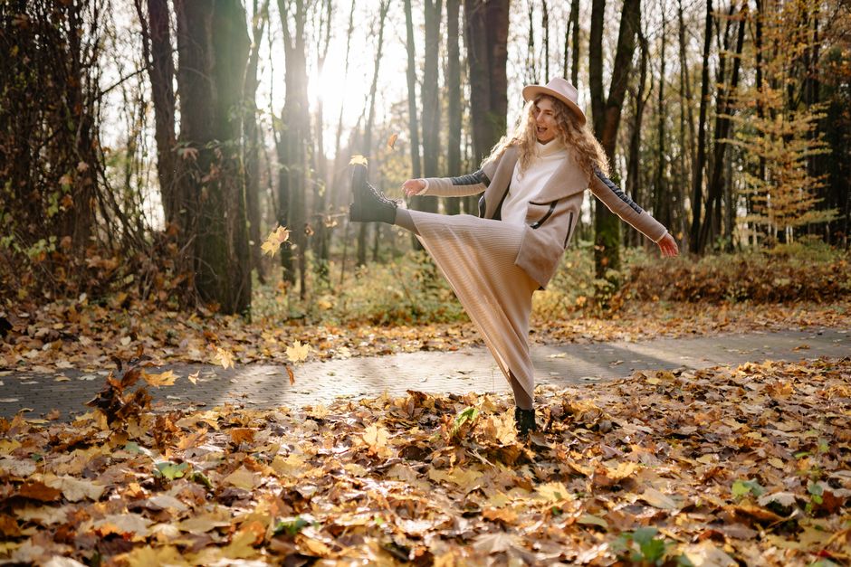 A happy woman with curly hair kicks leaves on an autumn day in the park.
