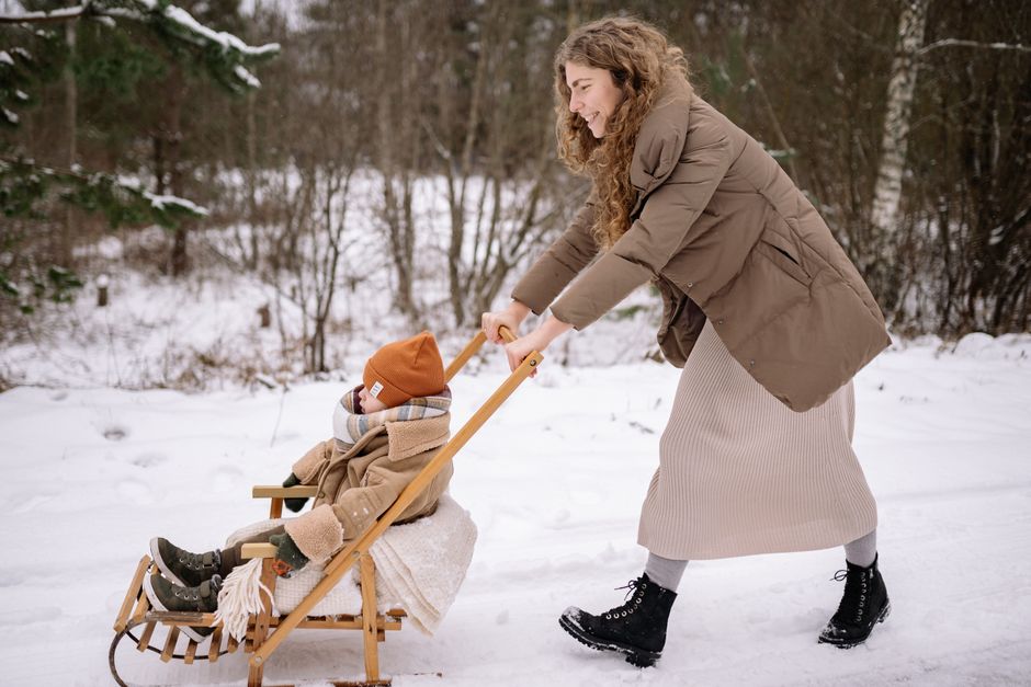 A mother pushing her child on a wooden sled in a snowy outdoor setting.