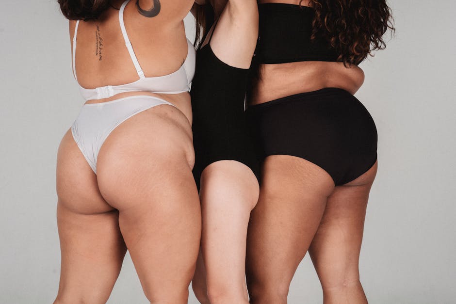 Back view of skinny female standing between plus size women in white and black underwear on white background in studio