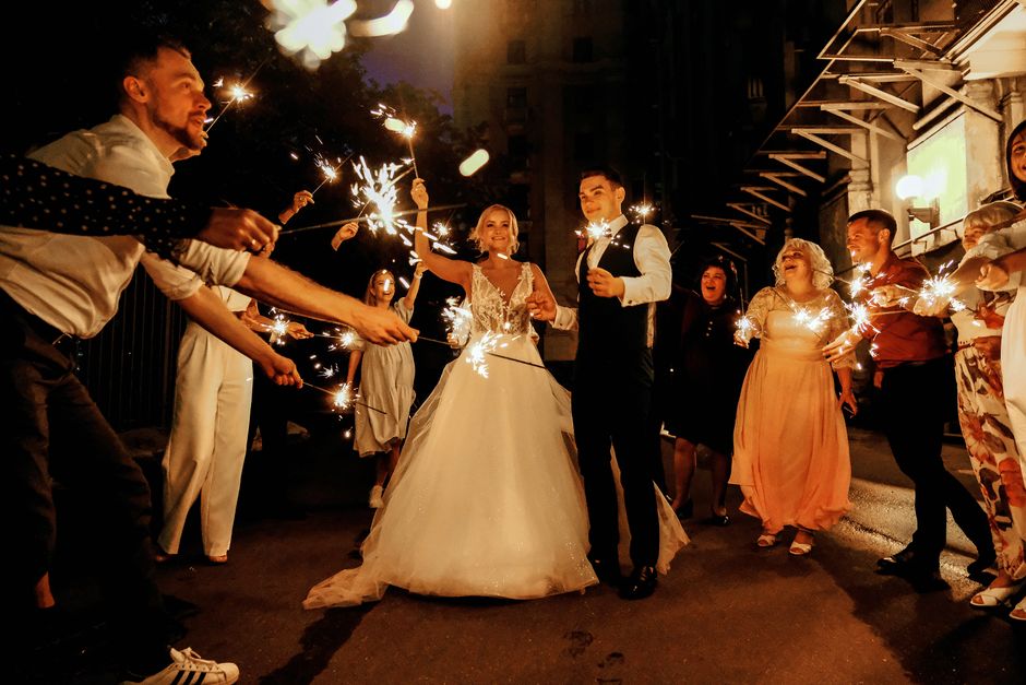 Full body of joyful young bride and groom with sparklers in hands walking near happy guests during wedding celebration in evening