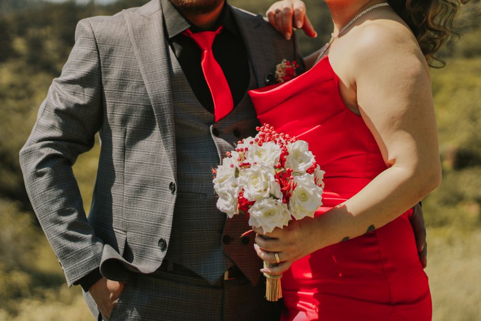 A couple elegantly dressed, the woman holding a bouquet, poses outdoors in a sunny setting.